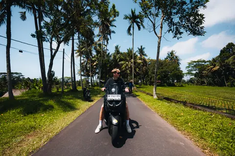 Malakai riding a scooter down a palm-lined road between rice fields