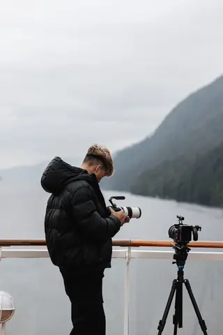 Malakai at a ferry rail with a camera on a tripod beside him, a fjord-like shoreline behind