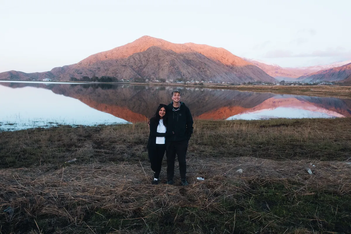 Malakai and Maddie standing together in front of a still lake that mirrors the mountains