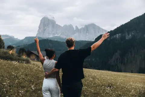 The two of them with their arms up in a wildflower meadow, a huge rock peak ahead